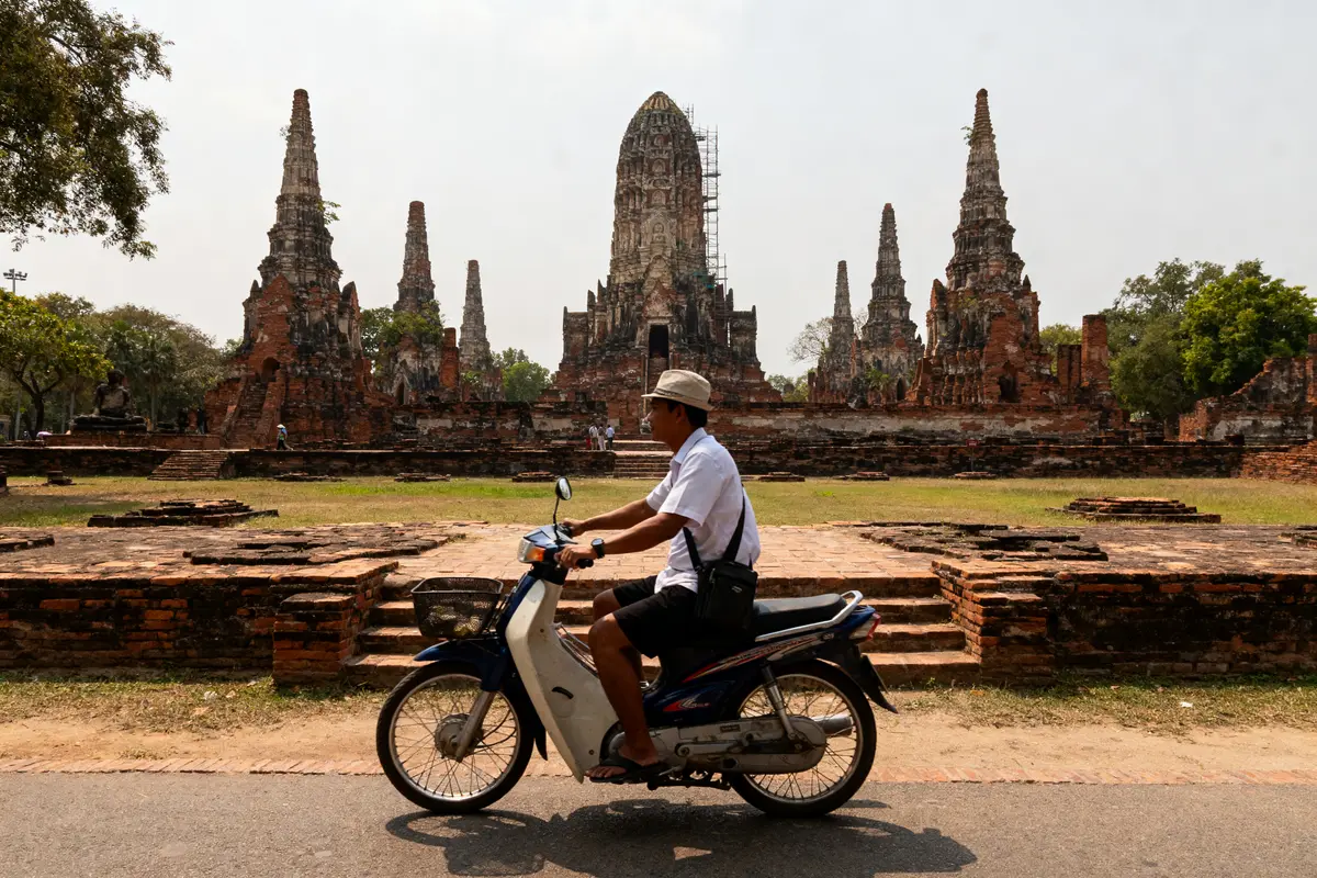 Homme à scooter devant temples anciens en briques, ciel clair, arbres verdoyants, journée ensoleillée.