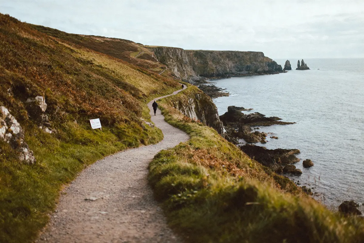 Sentier côtier bordant falaises verdoyantes, longeant la mer, avec deux promeneurs, sous un ciel nuageux.