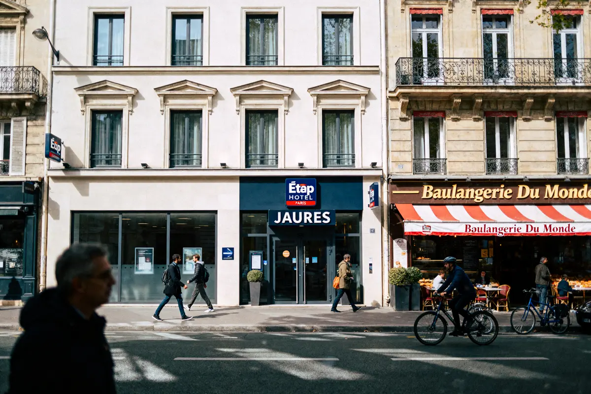 Façade de l'hôtel Jaurès à Paris, à côté d'une boulangerie, avec passants et cyclistes sur le trottoir.
