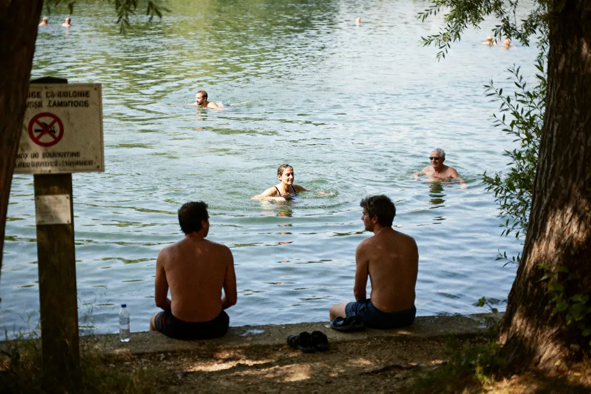 Personnes nageant dans un lac, deux hommes assis au bord, panneau de baignade réglementée visible.
